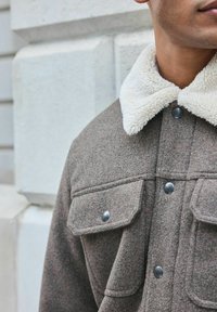 Braune Wollmischung Jacke mit einem beigen Kunstpelzkragen, zwei Brusttaschen und silbernen Druckknöpfen. Strukturierter Stoffdetails sichtbar.