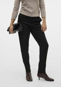 Pantalons noirs ajustés avec une coupe droite, fabriqués dans un tissu lisse, associés à des bottines brunes. Un sac à main noir est tenu dans une main.