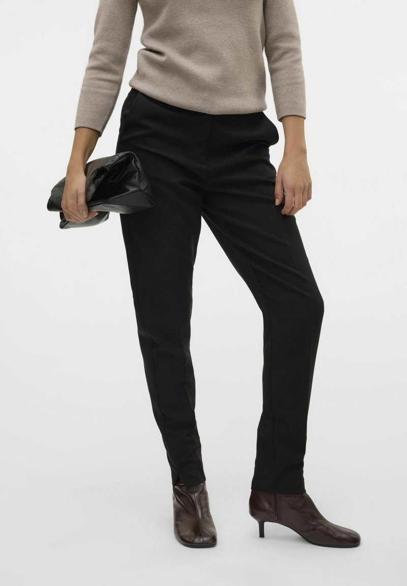 Pantalons noirs ajustés avec une coupe droite, fabriqués dans un tissu lisse, associés à des bottines brunes. Un sac à main noir est tenu dans une main.