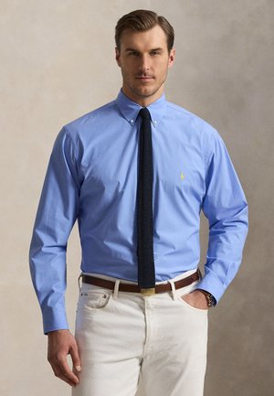 Mann trägt blaues Hemd mit Knöpfen, schwarze Strickkrawatte und weiße Hose, steht mit einer Hand in der Tasche vor neutralem Hintergrund.