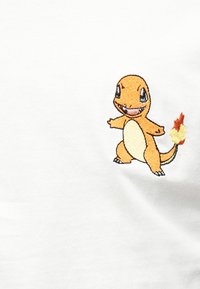 Μικρό κεντητό πορτοκαλί δεινοσαυράκι με φλόγα στην ουρά σε λευκό ύφασμα.