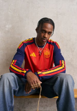 Rot und marineblaues Langarmtrikot mit gelben und roten Streifen an den Ärmeln, featuring ein spanisches Wappen und das Adidas-Logo. Kombiniert mit Denim-Hosen.