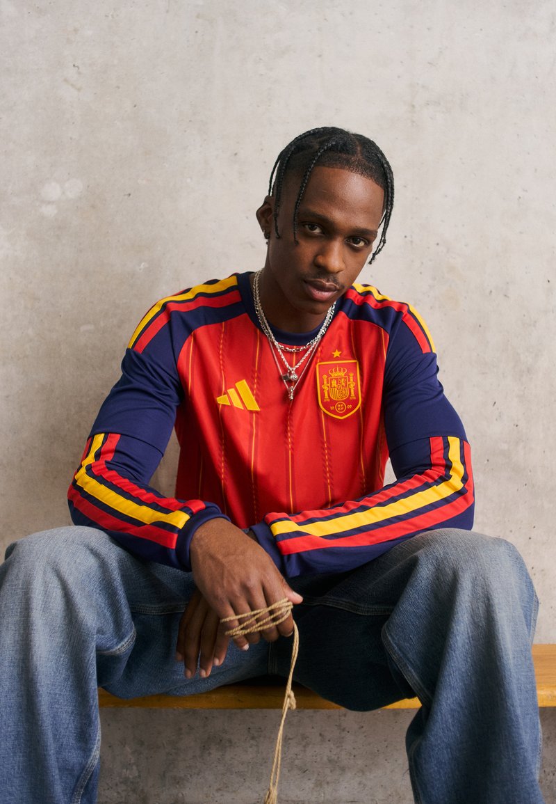 Κόκκινο και ναυτικό μπλε μακρυμάνικο τζέρσεϊ με κίτρινες και κόκκινες ρίγες στα μανίκια, διαθέτοντας ισπανικό έμβλημα και λογότυπο Adidas. Συνδυασμένο με τζιν παντελόνι.