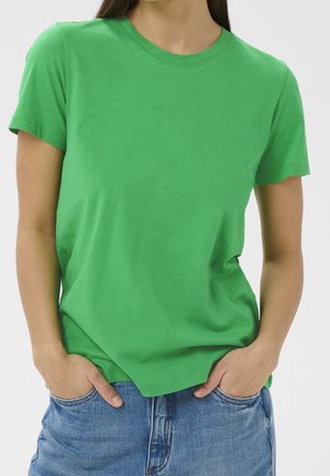 Personne portant un t-shirt vert uni à manches courtes et un jean bleu, les mains dans les poches, torse visible sur un fond uni.