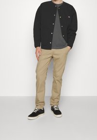 Man wearing black buttoned jacket over gray shirt, beige pants, and black sneakers against a plain white background.