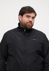Giacca leggera nera con colletto alto, chiusura con zip, tasche laterali e una piccola patch con logo sul petto. Texture liscia, design casual.