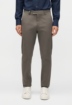 Άνδρας που φοράει ελαιοπράσινο παντελόνι στενής γραμμής, μπλε σκούρο πουκάμισο με κουμπιά και λευκά αθλητικά με κορδόνια, στέκεται μπροστά σε μονόχρωμο φόντο.