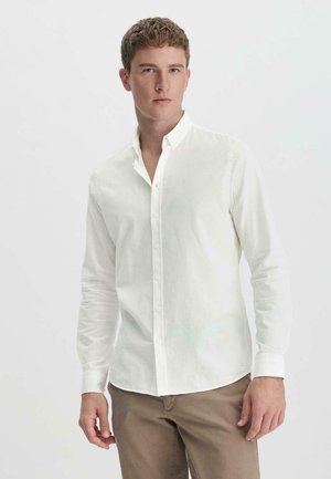 Camicia bianca a maniche lunghe con colletto button-down, design slim fit e texture morbida. Indossata con pantaloni beige, che mettono in risalto uno stile minimalista.