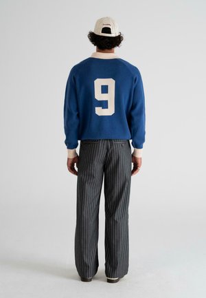 Man wearing a blue sweater with a white number 9 on the back, striped gray pants, and a white cap facing away from the camera.