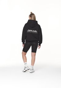 Felpa nera in cotone con un grande testo grafico sulla schiena, abbinata a pantaloni ciclistici neri e sneakers bianche, su uno sfondo chiaro.