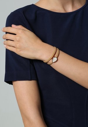 Bracelet en caoutchouc beige clair avec un accent argenté en forme de goutte, empilé avec un bracelet rond marron, porté au poignet contre un haut bleu marine.