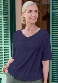 Marineblaue, leichte Bluse mit einem drapierten Ausschnitt und kurzen Ärmeln, mit lässigem Schnitt. Kombiniert mit hellgrünen Hosen.