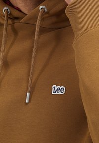 Ruskea huppari pehmeällä tekstuurilla, jossa valkoinen Lee-logo kirjailuna. Mukana kiristysnauha hopeisilla päätteillä.