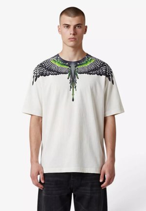 Giovane uomo che indossa una t-shirt bianca con un motivo di ala di uccello nero e verde intorno al collo, abbinata a pantaloni neri.