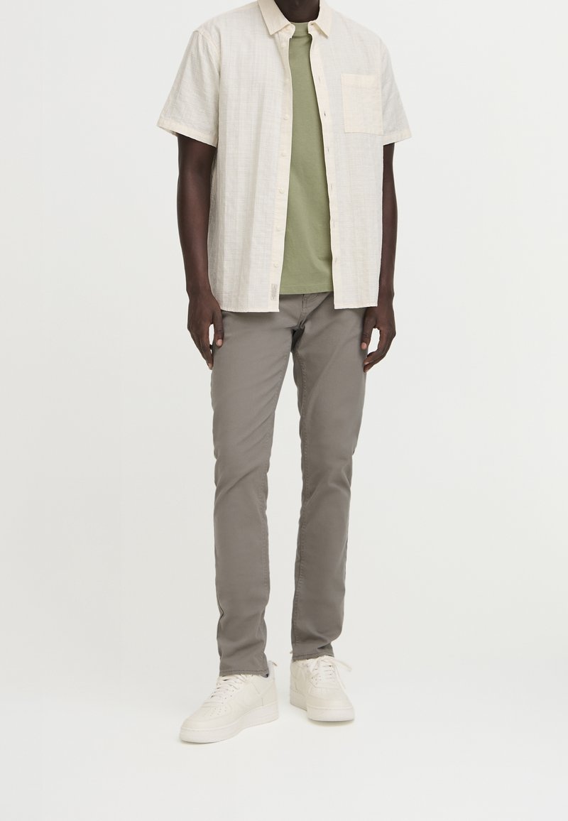 Homme portant une chemise crème à manches courtes boutonnée par-dessus un t-shirt vert olive, un pantalon gris slim et des baskets blanches, debout devant un fond uni.