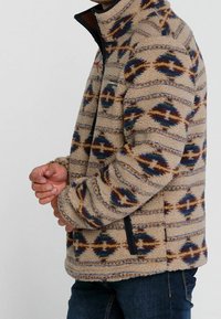 Fleecejack met een beige kleur en een patronen van blauwe en bruine diamanten. Voorzien van een hoge kraag en zijzakken met rits.