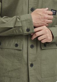 Khaki groene jas van een gestructureerde stof, met zwarte knopen, een logo patch en twee voorzakken met kleppen.