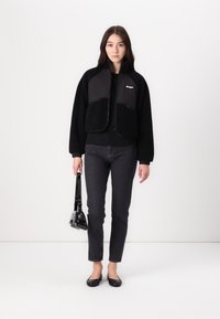 Giacca nera corta con maniche in fleece, chiusura con zip e due tasche frontali. Il modello indossa jeans grigi e tiene una piccola borsa nera.