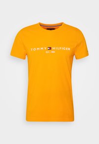 Camiseta de algodón naranja con cuello redondo, que presenta el texto blanco "TOMMY HILFIGER EST. 1985" y un acento de logo en el centro. Ajuste estándar.