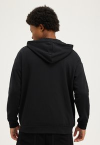 Lyhyttukkainen kiharatukkainen henkilö, joka on kuvattu takaa, pukeutuneena yksiväriseen mustaan huppariin ja mustiin housuihin vaaleaa taustaa vasten.