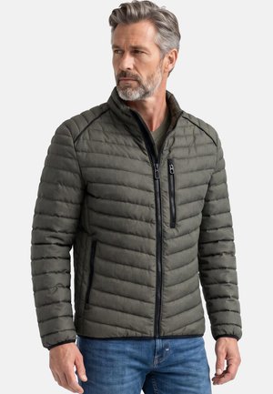 Mand i midten af 40’erne med gråt hår og skæg iført en grøn quiltet jakke og blå jeans, der kigger til venstre mod en ensfarvet baggrund.