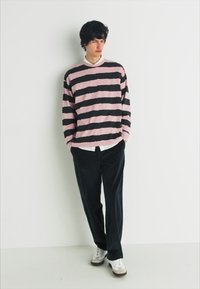 Pull à rayures à manches longues en rose et noir, superposé sur une chemise blanche à col, associé à un pantalon sombre et des baskets blanches.