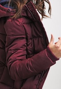 Bordowa kurtka puchowa o fakturowanym wykończeniu, z zamkiem błyskawicznym, ściągaczami na mankietach i kapturem. Detale przeszyć widoczne wzdłuż szwów.