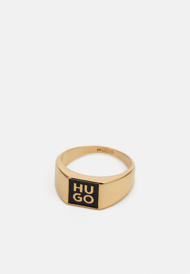 Anello in tono dorato con un accento rettangolare nero su cui è scritto "HUGO" in testo bianco; superficie liscia, finitura lucida e design a banda arrotondata.