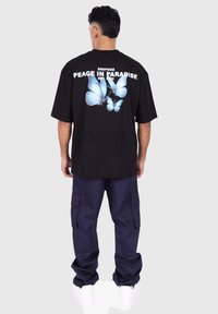 Black oversized t-shirt featuring blue butterfly graphic and text on the back, paired with navy cargo pants and white sneakers.