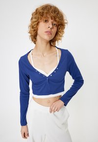 Geknipte, langmouw top in blauwe ribbed stof met een diepe V-hals, voorzien van decoratieve knopen en een contrasterende witte rand aan de zoom.