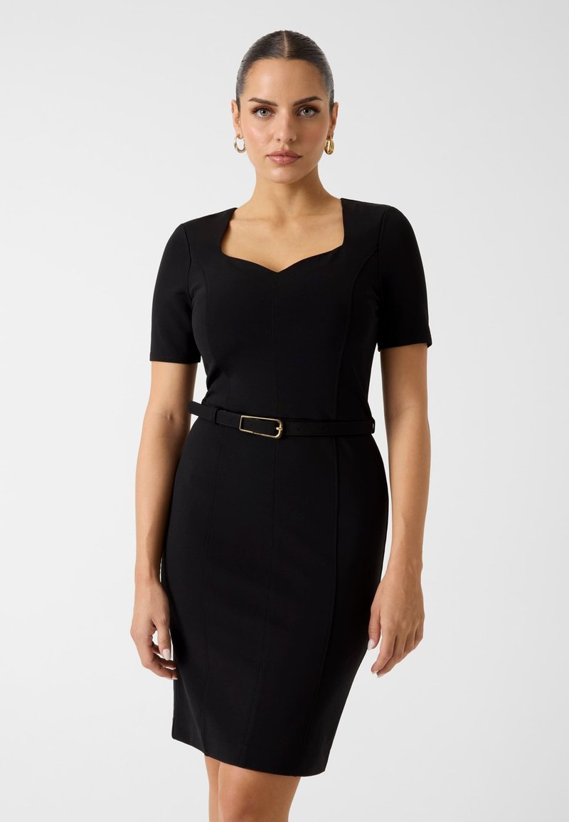 Femme aux cheveux foncés coiffés en un chignon lisse, portant une robe noire ajustée à manches courtes et une fine ceinture noire avec une boucle dorée.