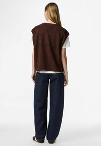 Brun texturerad ärmlös topp över en vit skjorta, matchad med mörkblå vida jeans, sedd bakifrån.