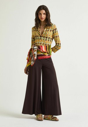 Donna che indossa una camicia gialla senape a fantasia, fascia multicolore annodata in vita, pantaloni neri a gamba larga e sandali coordinati con fantasia.