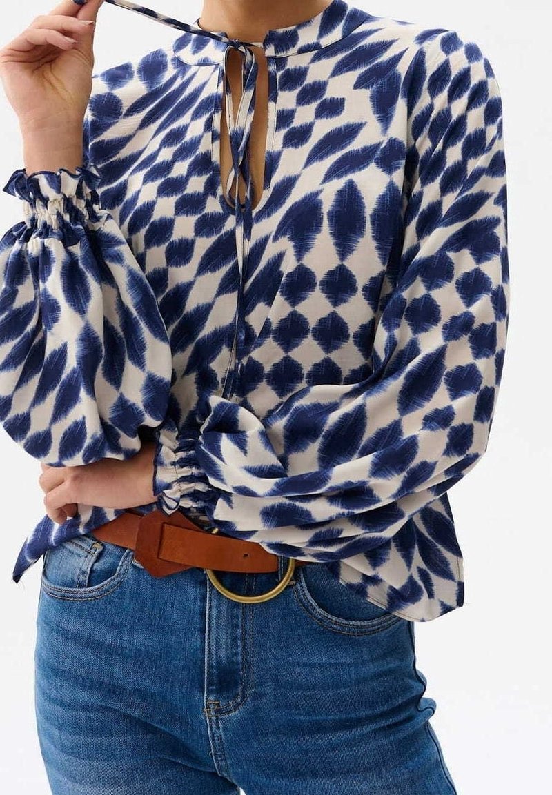 Femme portant un chemisier bleu et blanc à motifs avec de longues manches bouffantes, col noué, ceinture marron et jean bleu, main près du cou.