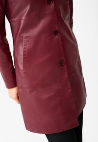 Burgundy leather coat featuring a smooth texture, double-breasted button closure, and side pocket. Tailored cut with a straight fit.