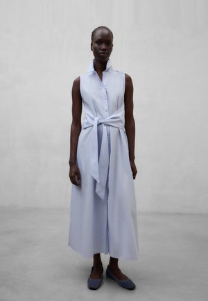 Mujer con vestido sin mangas azul claro abotonado y con la cintura anudada, de pie contra un fondo gris liso, vistiendo zapatos planos azules.