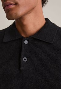 Polo preto texturizado com um tecido macio e canelado. Apresenta um colarinho clássico e dois botões pretos na gola.