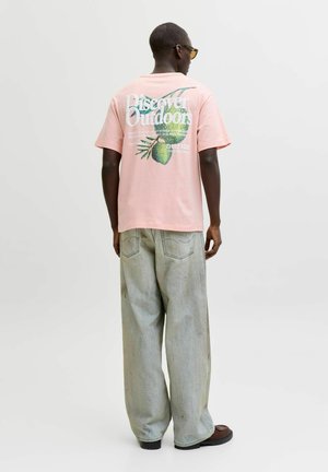 Mężczyzna w różowym T-shircie z napisem „Discover Outdoors” i grafiką ananasa na plecach, jasnych szerokich dżinsach i brązowych butach, stojący tyłem.