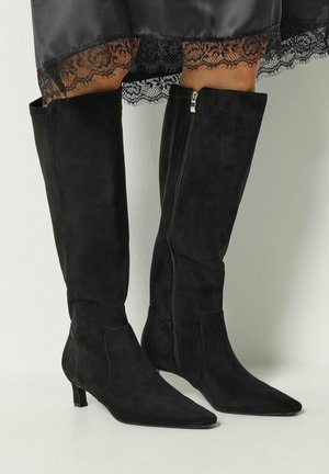 Femme portant des bottes hautes en daim noir à bouts pointus et à talons plats, assorties à une jupe noire ornée de dentelle.
