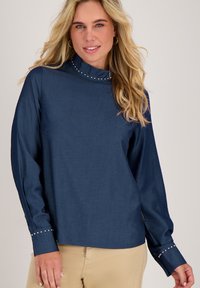 Donkerblauw langmouw shirt met hoge kraag en decoratieve zilveren studs langs de halslijn en manchetten. Glad stof, ontspannen pasvorm.