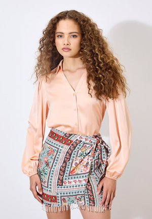 Blouse rose clair avec un devant à boutons et des manches bouffantes, associée à une mini-jupe à motifs présentant des designs floraux et géométriques, avec des franges.