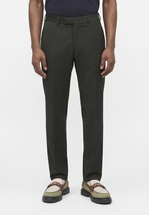 Mand iført tætsiddende mørkegrønne bukser og tofarvede loafers med guldkædedetalje, stående mod en hvid baggrund.