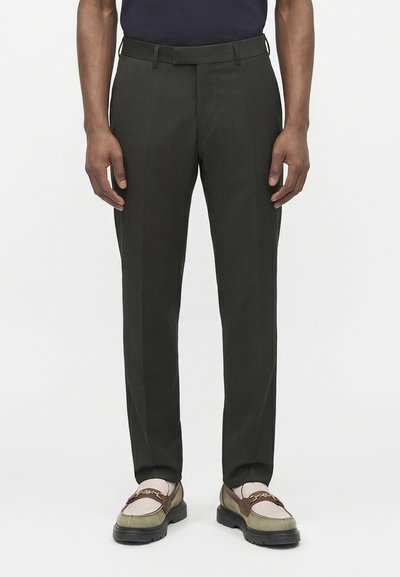 Mann trägt eng anliegende dunkelgrüne Hose und zweifarbige Loafers mit goldener Kettendetail, steht vor weißem Hintergrund.