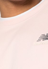Felpa rosa chiaro con colletto a coste e logo "new balance" ricamato in grigio scuro, realizzata in un tessuto morbido e texture.