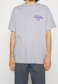 Mand iført en lys grå t-shirt med korte ærmer, som har lilla "Carhartt Carrosserie Wip" tekst på brystet, og mørke grå bukser.