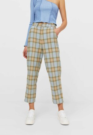 Mujer vistiendo pantalones cropped de cuadros de talle alto en beige y azul con zapatillas blancas y un top de punto azul claro de un solo hombro.