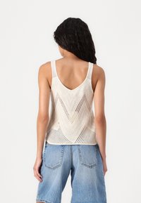 Knit sleeveless top in cream with a chevron pattern and open weave. Features wide straps and a deep back neckline. Paired with denim shorts.