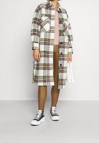 Cappotto di lana scozzese nei toni del bianco, rosso, giallo e nero. Presenta ampie tasche sul petto, chiusura con bottoni e spacco laterale. Indossato con leggings color sabbia e sneakers.