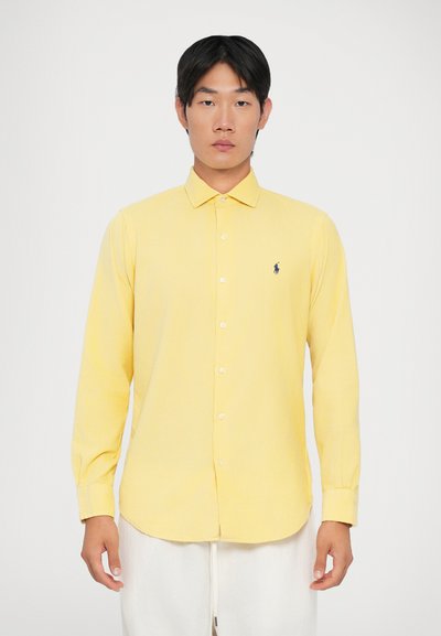 Camisa amarilla de manga larga con botones, de textura suave y cuello puntiagudo, que presenta un pequeño logo azul en el lado izquierdo del pecho.