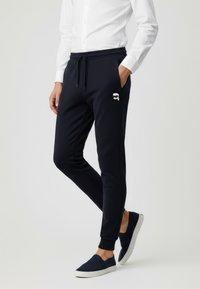 Navyblaue Jogginghosen aus weichem Stoff mit Seitentaschen und elastischen Bündchen. Mit einem weißen Logo auf dem Oberschenkel.